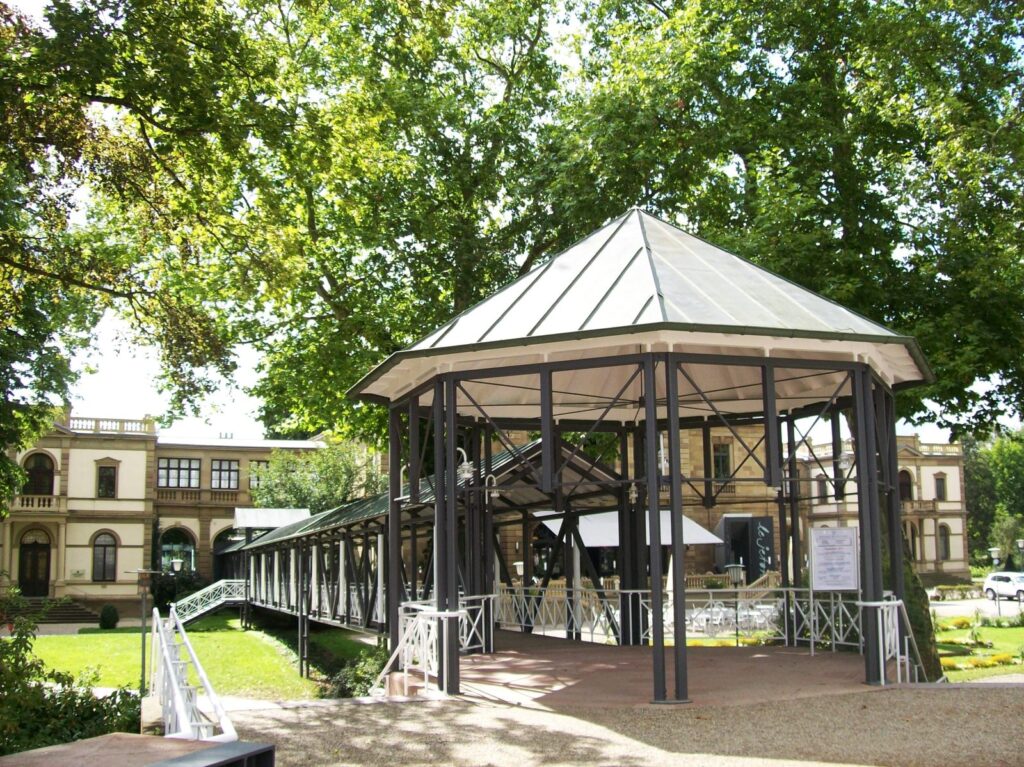 Offener Pavillon mit Zeltdach und angrenzender Wandelgang in einem Kurpark