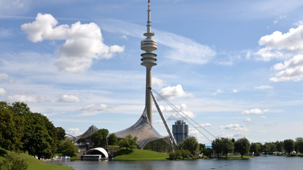 Olympiaturm über dem Olympiasee mit Zeltdach und Uferwiese im Sommer