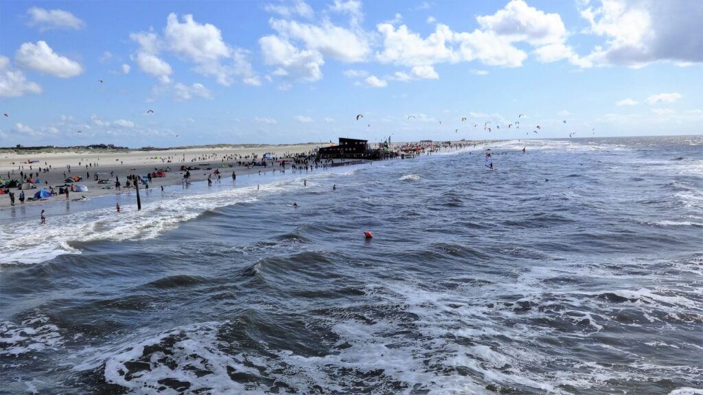 Breiter Sandstrand von St. Peter-Ording mit Pfahlbau, Brandung und vielen Kite-Schirmen