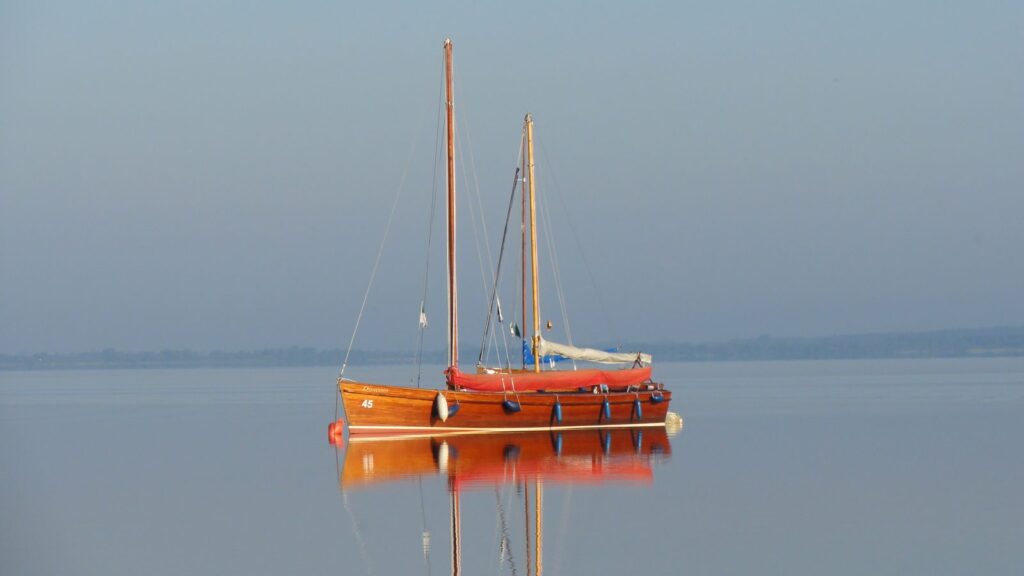 Hölzernes Auswandererboot mit zwei Masten liegt bei ruhigem Wasser vor Anker