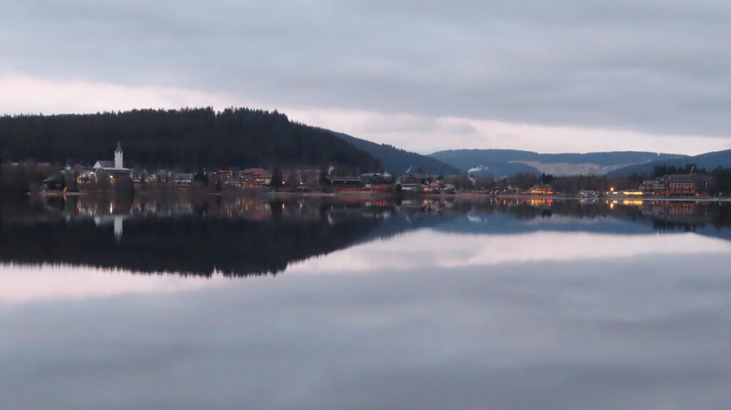 Blick vom Seerundweg über den Titisee auf den Ort mit Kirchturm und bewaldeten Hängen