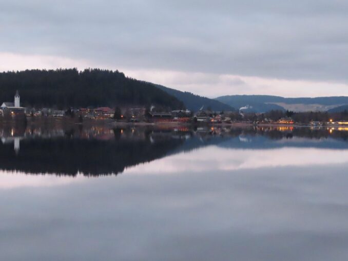 Blick vom Seerundweg über den Titisee auf den Ort mit Kirchturm und bewaldeten Hängen