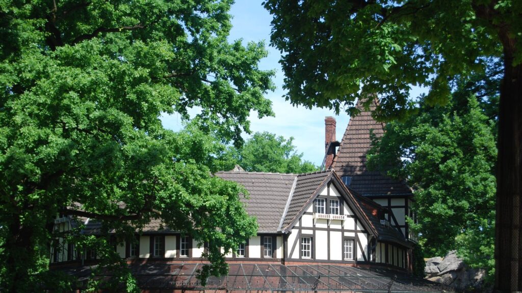 Historisches Fachwerkgebäude im Zoo Berlin hinter alten Bäumen mit Voliere davor
